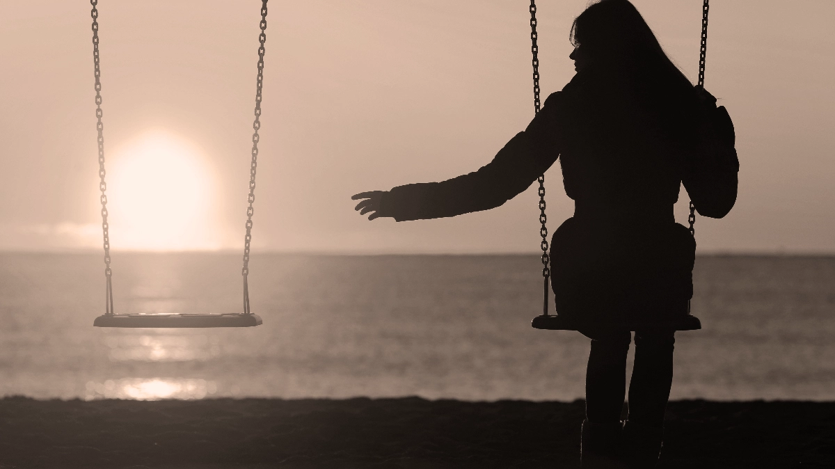 Mujer sentada en un columpio frente al mar, extendiendo su mano hacia un columpio vacío adyacente mientras el sol del atardecer se pone en el horizonte, simbolizando el proceso de superar el duelo.
